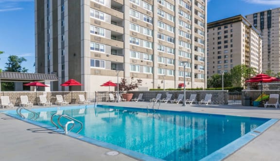 a swimming pool with chaise lounge chairs and umbrellas in front of a tall building at The Ivanhoe Apartments, Hackensack, New Jersey