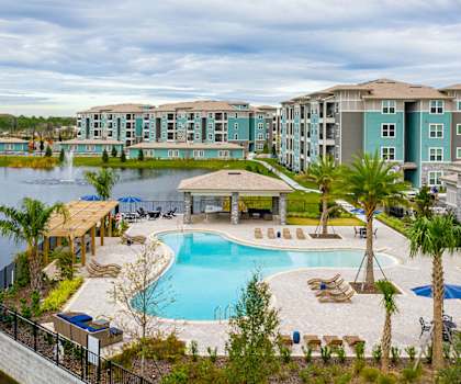 A large swimming pool surrounded by palm trees and lounge chairs in front of apartment buildings.
