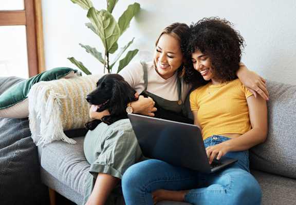 two women sitting on a couch with a dog and a laptop