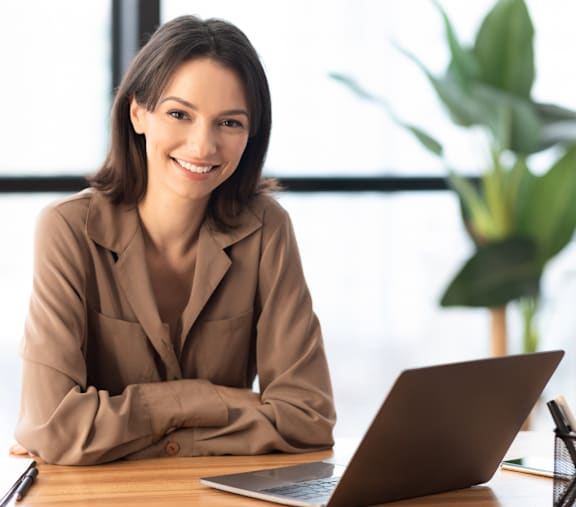 A woman in a brown blazer is sitting at a desk with a laptop.