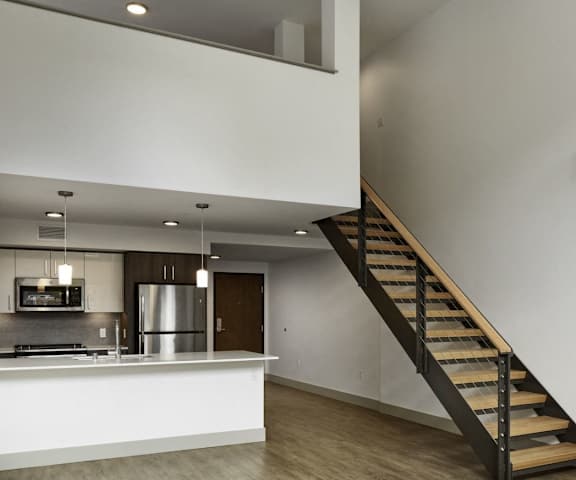 The view from the front entrance showing a kitchen with stainless steel appliances, white cabinets, and large white counters,, hallway with closets and overhead lighting, and carpeted stairs leading to the top floor at Uptown at Kirkland Urban. 