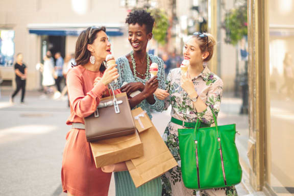 Three women are standing outside a store, one of them is holding a green bag.