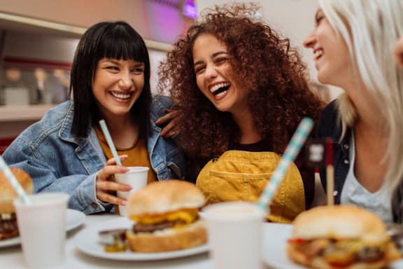 Three women are laughing and eating burgers at a table.