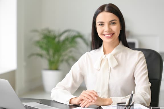 A woman in a white blouse is sitting at a desk with a laptop.