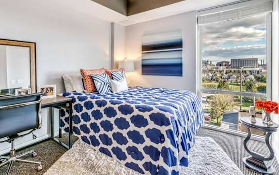 A bedroom with a blue and white bedspread, a desk with a chair, and a view of the city.