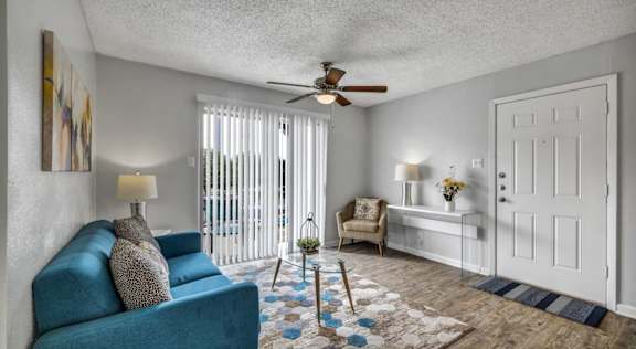 A blue couch sits in a living room with a ceiling fan and a window with blinds.