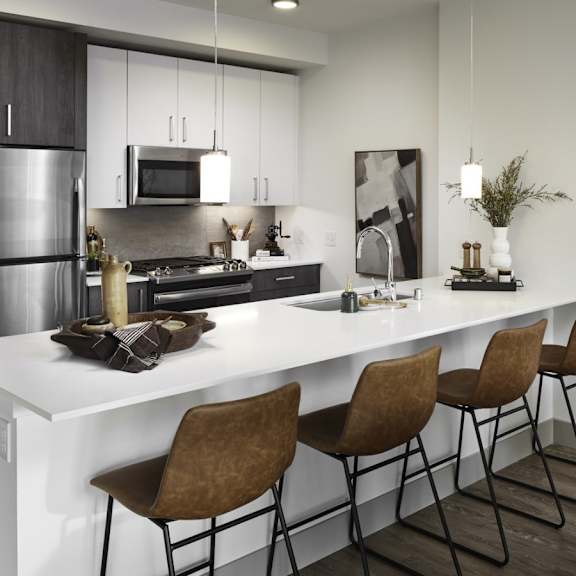 A spacious, modern kitchen with stainless steel appliances, kitchen utensils, overhead lighting, light countertops with lots of room, a bar counter with barstools, and large wooden cabinets at Uptown at Kirkland Urban. 