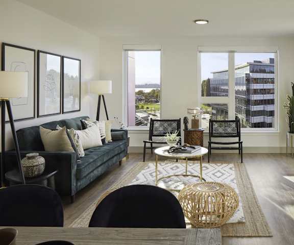 The spacious living room with a slate gray couch, black and white lamps, white throw pillows, large windows, a ceiling fan, wall art, plush chairs, coffee table, decorations, large windows, a TV, and glass doors to the balcony at Uptown at Kirkland Urban.
