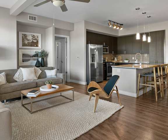 The spacious living room with a gray couch, white plush chairs, one navy chair, coffee table, overhead lighting with a ceiling fan, decorations, a TV, and view of the dining room with a table, chairs, and place settings for four, and the kitchen at Skywater at Tempe Town Lake. 