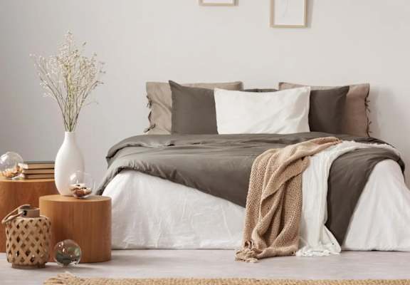 A bed with a grey comforter and pillows is in the center of a room with a white wall and a white floor.