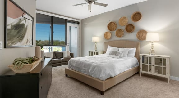 A spacious bedroom with a queen-size bed with a white bedspread, a dresser, a dark gray couch, overhead lighting with a ceiling fan, wall art, lamps, decorations, window, door to the balcony, and nightstands with lamps at Skywater at Tempe Town Lake Apartment Homes. 