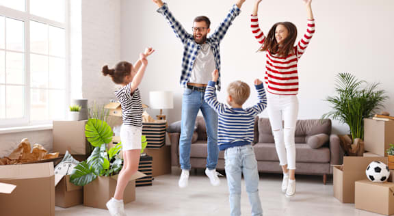 A family is excitedly jumping in their new home surrounded by moving boxes.