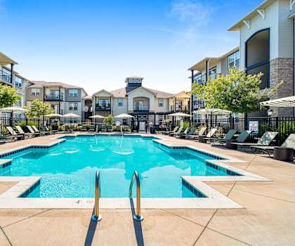 A large swimming pool surrounded by lounge chairs and umbrellas in front of apartment buildings.