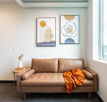 A tan couch with an orange throw is in front of a window with two framed pictures on the wall.