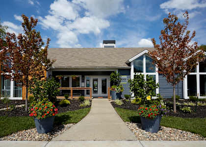 A house with a grey roof and a white door is surrounded by trees and flower pots.