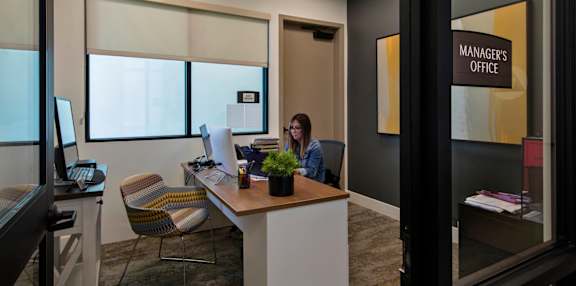 A woman is sitting at a desk in an office with a sign that says "Manager's Office" on the wall behind her.