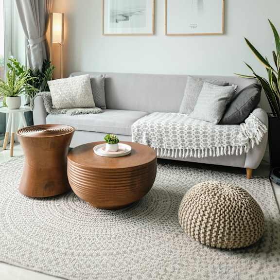 A living room with a grey couch, a brown vase, a round knitted ottoman and two framed pictures on the wall.