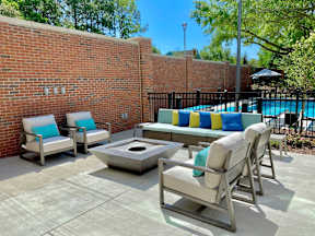 An outdoor community lounge with 4 comfortable chairs, a sofa, and a fire pit near a gated swimming pool.