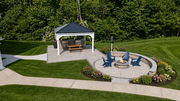 A gazebo with a fire pit and chairs is surrounded by a garden.