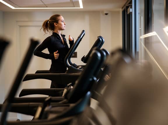 A woman in a black top is running on a treadmill in a gym. at Trio Newton Apartments, Newton, Massachusetts