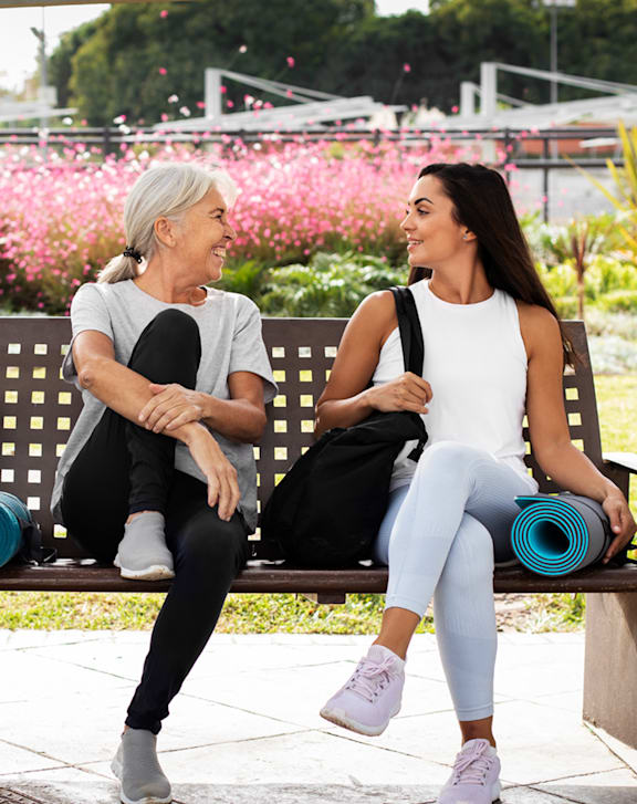 Two women sitting on a bench with one holding a yoga mat.