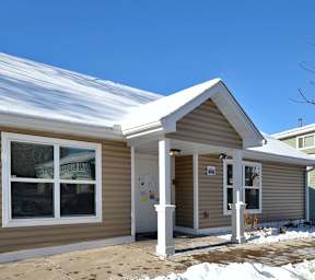 A small house with a brown siding and a white trim is covered in snow.