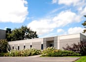 Thumbnail 1 of 3 - A modern building with a grey facade surrounded by greenery and a clear blue sky.