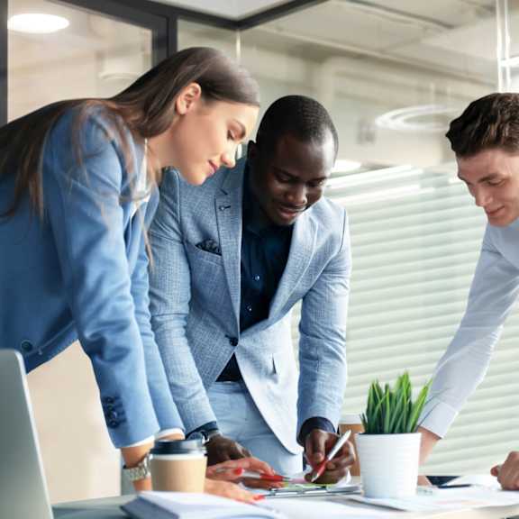 Three business people in suits are gathered around a table looking at documents.