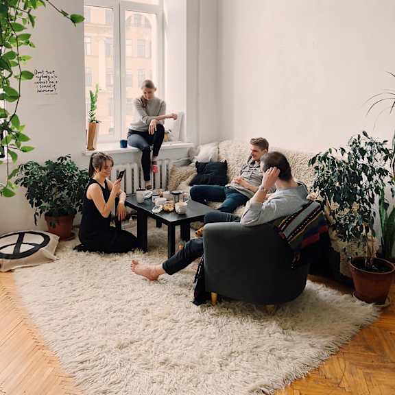 A group of people are sitting in a living room with a white rug and a large window.