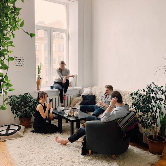A group of people are sitting in a living room with a white rug and a large window.