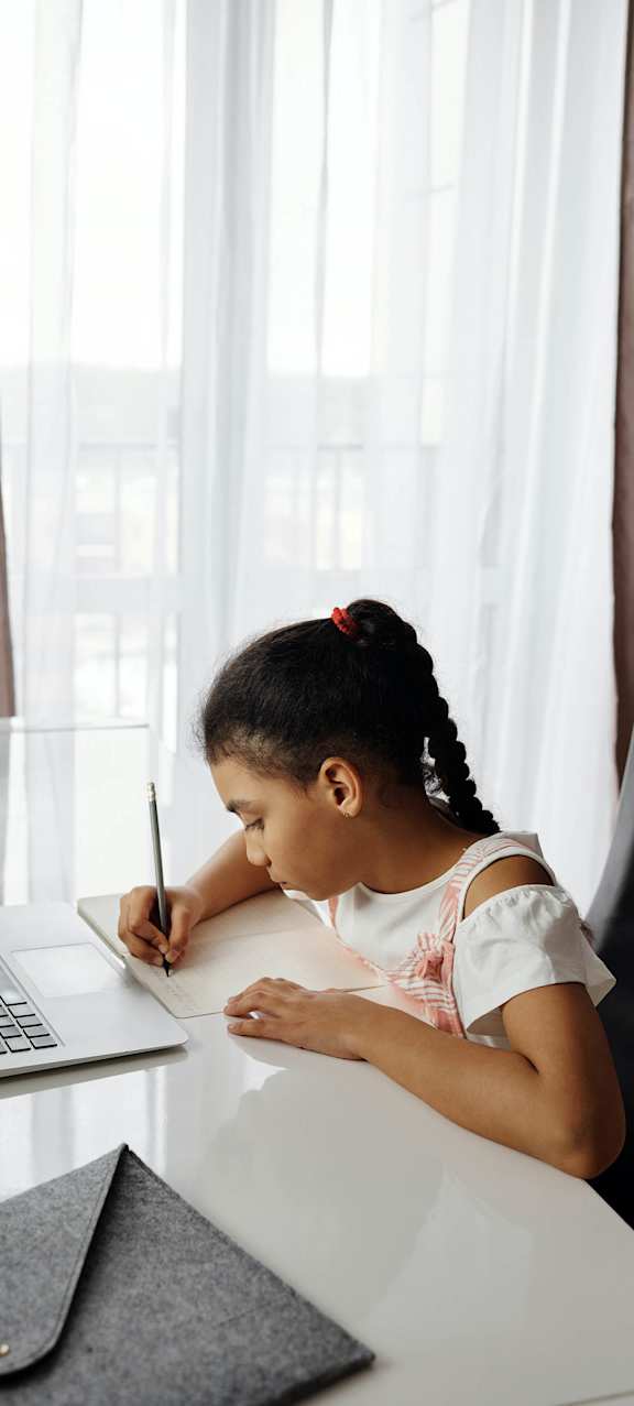 A girl is writing on a notepad while sitting at a desk with a laptop.