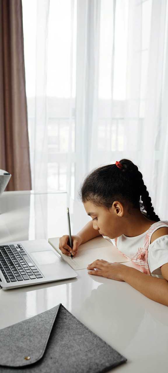 A girl is writing on a notepad while sitting at a desk with a laptop.