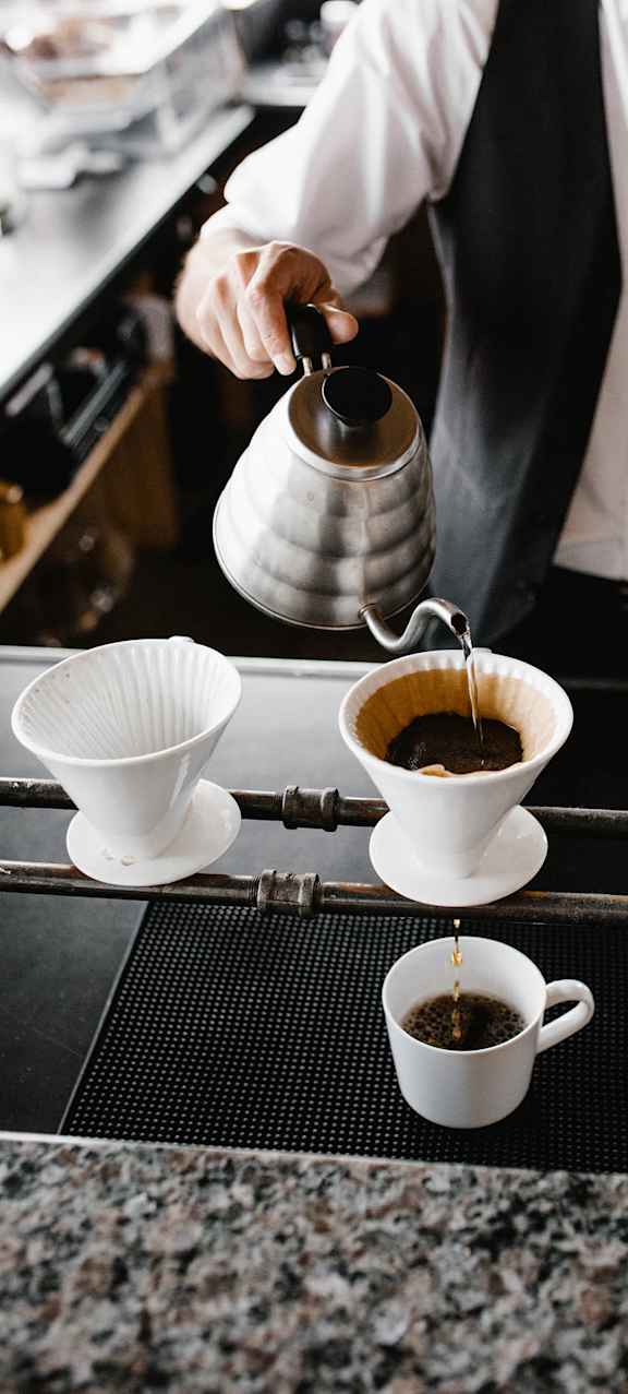 A person is pouring coffee from a silver pot into a white cup.
