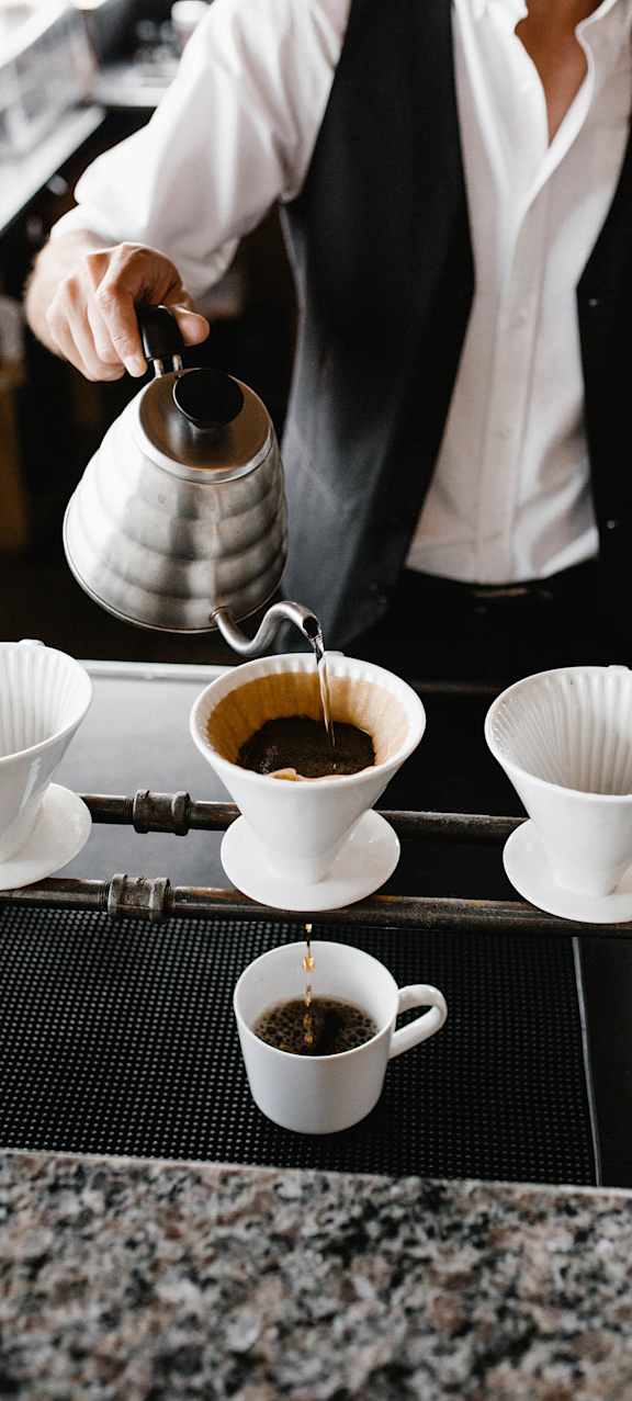 A person is pouring coffee from a silver pot into a white cup.