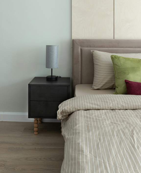 A lamp sits on a nightstand next to a bed with striped bedding.