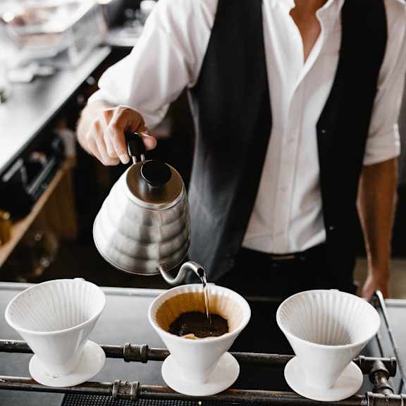 A person is pouring coffee from a silver pot into a white cup.