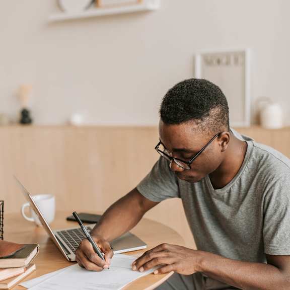 A man is writing on a piece of paper at a desk with a laptop and other items around him.