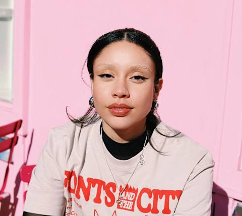 A woman wearing a pink shirt with the words "Fonts and City" on it is sitting in front of a pink wall.