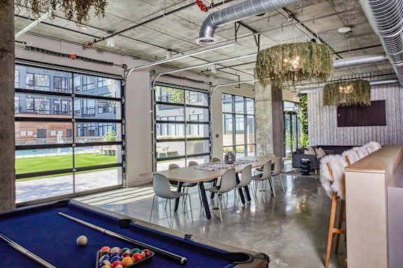 A pool table with a blue felt cloth and a set of pool balls in a room with a high ceiling and large windows.