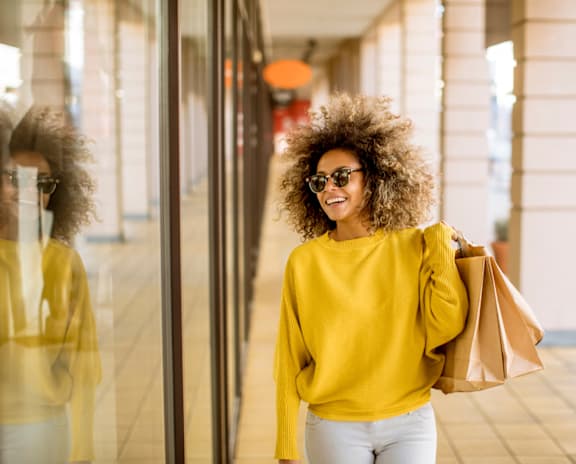 A woman with a yellow sweater and white pants is holding a brown paper bag while standing in front of a glass door.