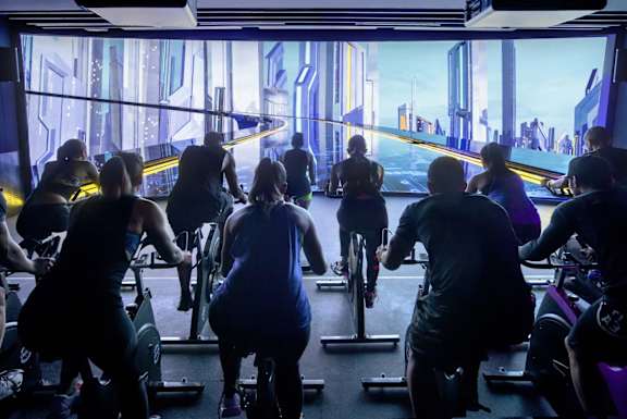A group of people are exercising on stationary bikes in a room with a futuristic cityscape displayed on the wall behind them.