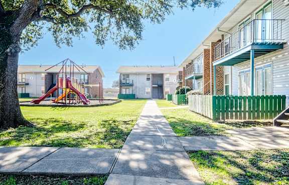 A playground with a slide is in the middle of a grassy area between two apartment buildings.