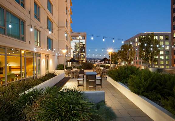 A rooftop patio with a table and chairs is illuminated by string lights at The Grand Apartments, Oakland, CA, 94612