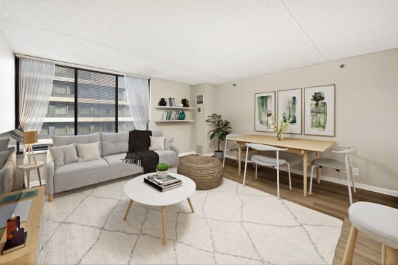 A living room with a grey sofa, a white coffee table, a wooden dining table and chairs, a rug, and a large window with a view of a building outside. at Lenox Club Apartments, Arlington, VA, 22202