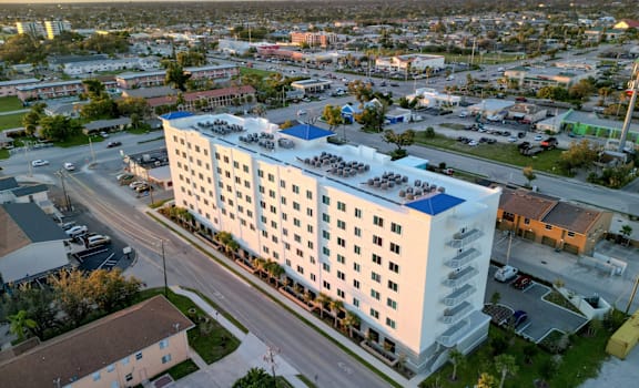 A large white building with a blue roof is surrounded by a parking lot and other buildings.