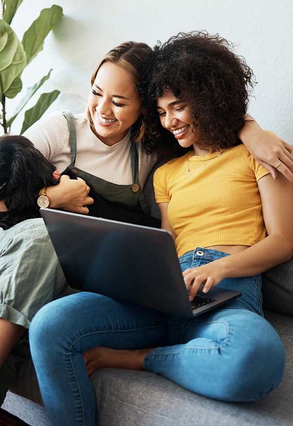 two women sitting on a couch with a laptop computer and a dog