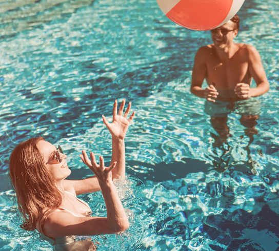 A woman reaching for a beach ball in a pool.