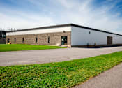 Thumbnail 2 of 4 - A large industrial building with a grey roof and a green lawn in front.
