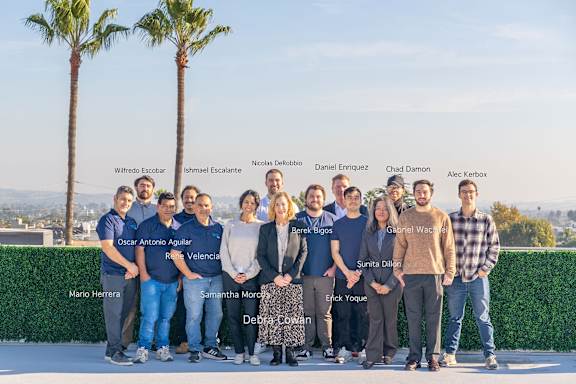 A group of people are standing in front of a hedge with palm trees in the background.