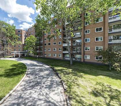 A tree-lined walkway leads through a grassy area between two apartment buildings.